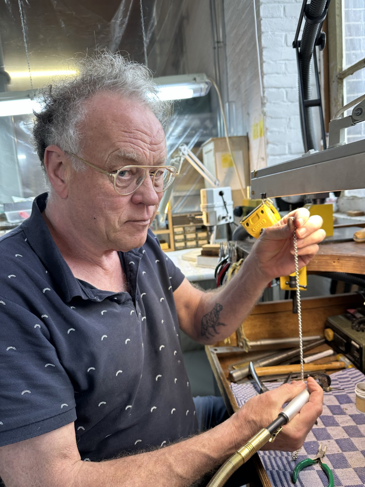 Een man met een bril werkt in een atelier aan een zilveren ketting met handgereedschap boven een werkbank met hamers en tangen.